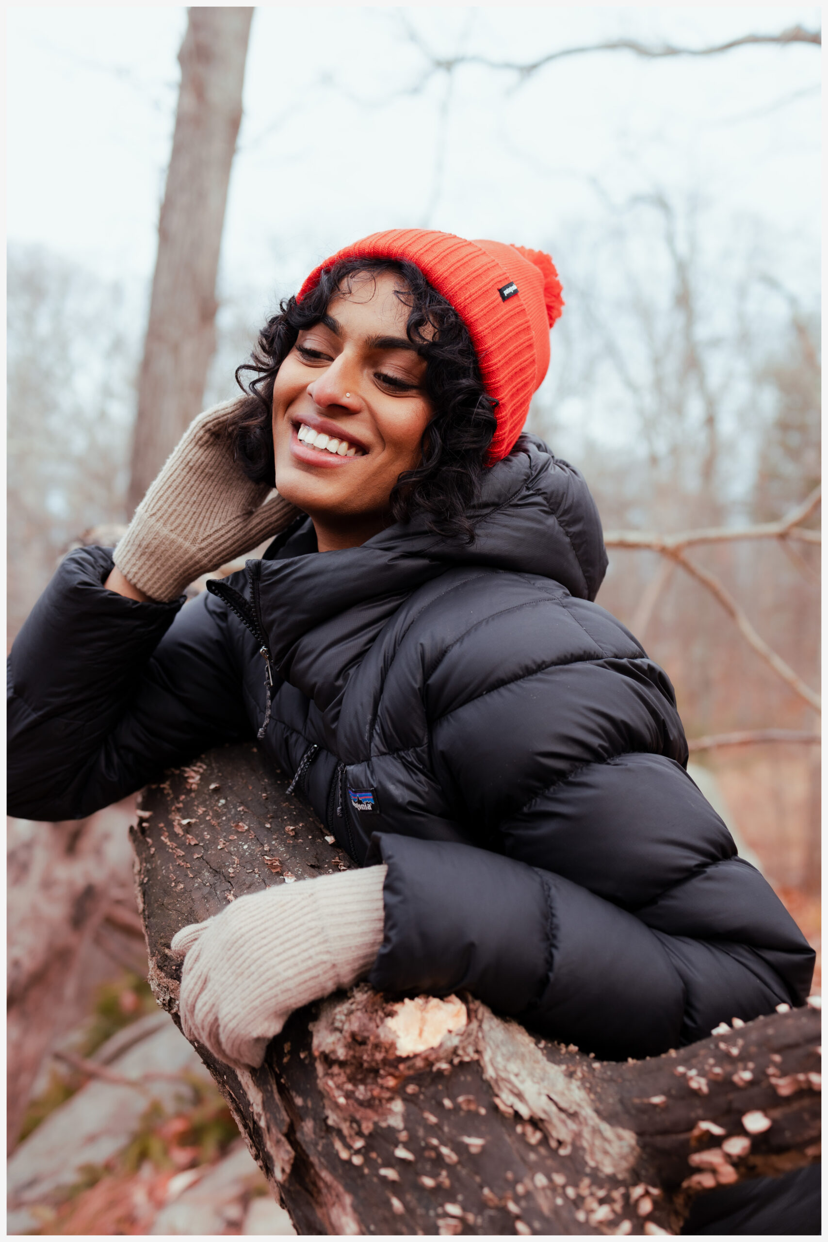 Woman in an orange beanie and mittens smiling, leaning on a fallen tree